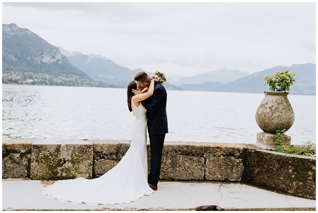 bride and groom hugging and kissing in front of mountains and lake in annecy france