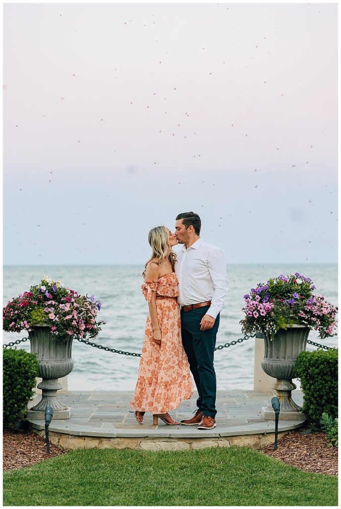 Woman in orange dress kisses man by water during Grosse Pointe Engagement Session