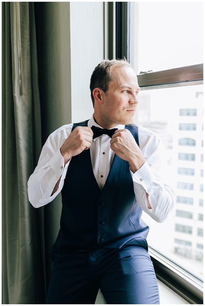 Man adjusts bowtie looking out window by Detroit Wedding Photographer.