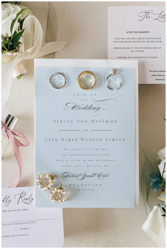 Three rings and wedding jewelry are on top of wedding stationery suite by by Detroit Wedding Photographer.
