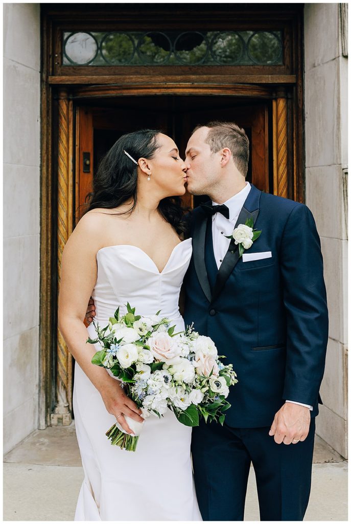 Newly married, husband and wife, share kiss outside building by Detroit Wedding Photographer.