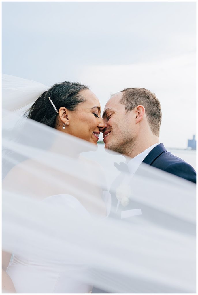 Noses touch and smiles are shared while veil flows in front of couple by by Detroit Wedding Photographer.