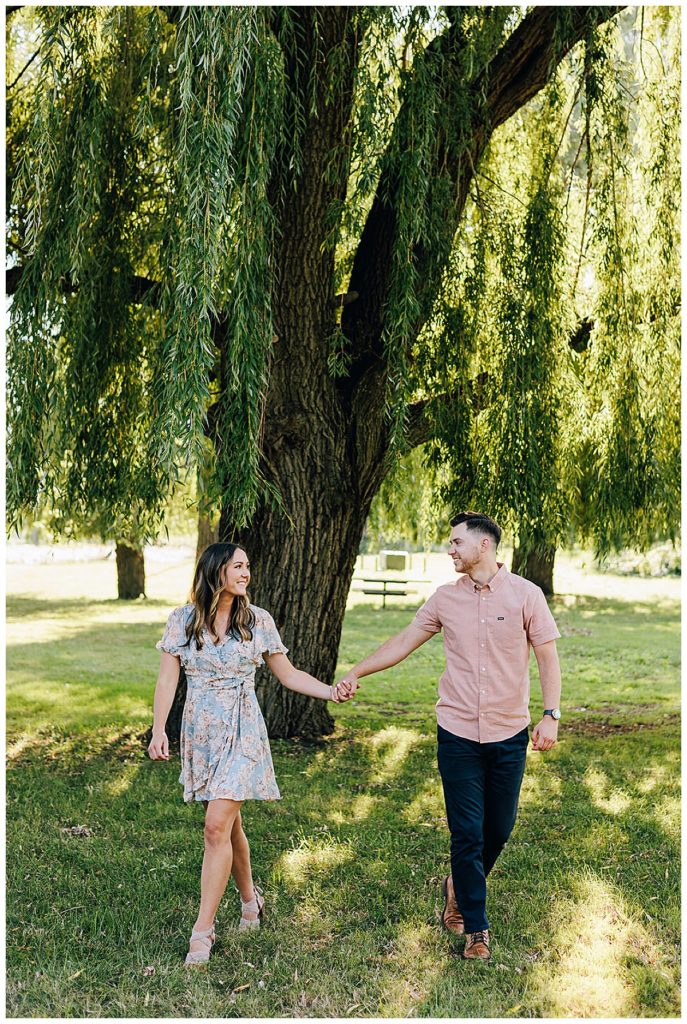 Man and woman walk hand in hand under willow tree for Belle Isle engagement