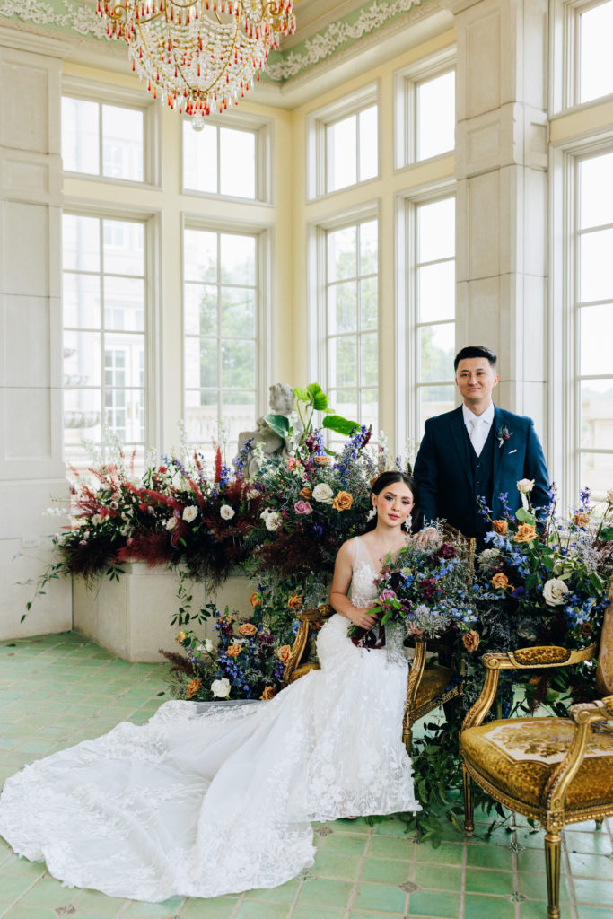bride in chair and groom standing next in front of floral installation for Kayla Bouren photography