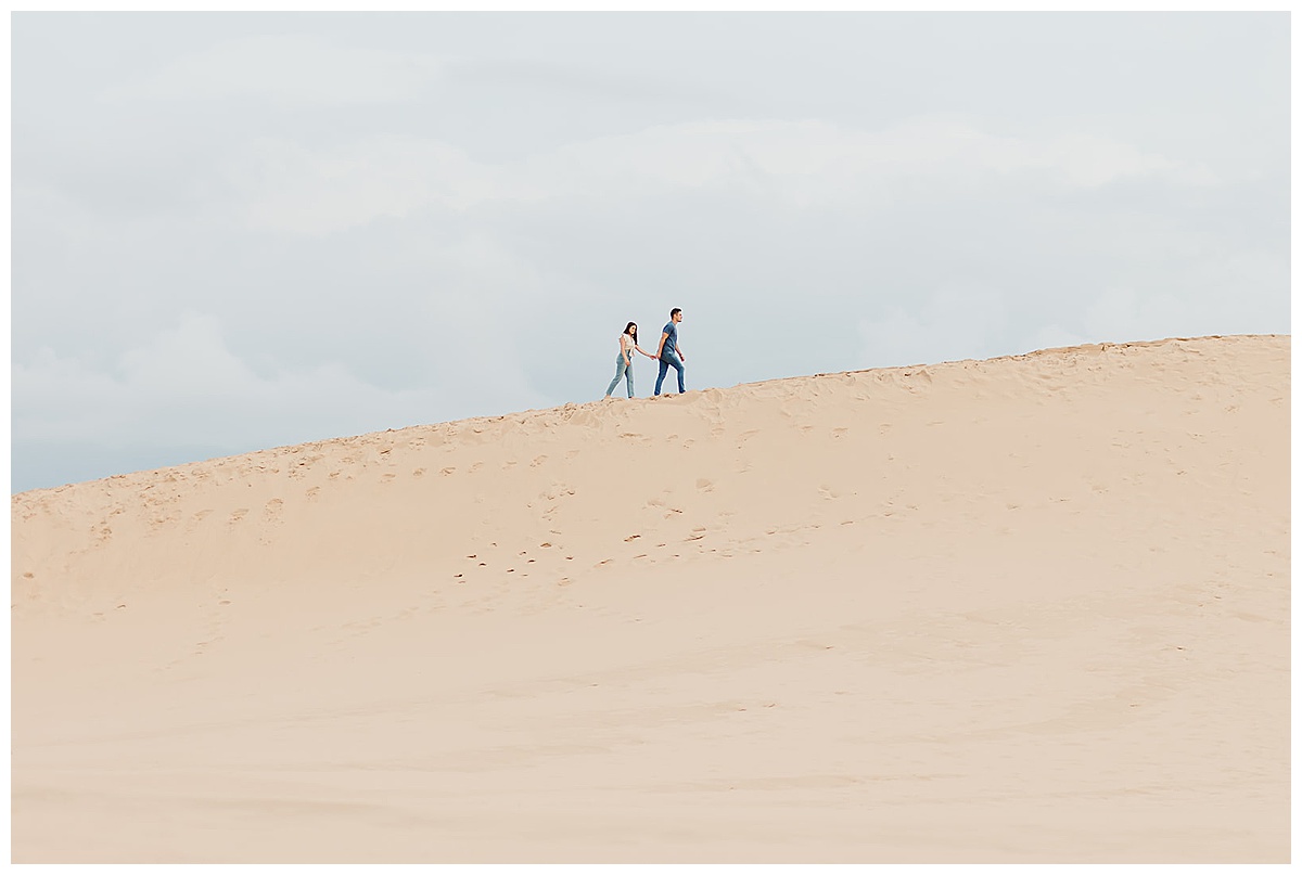 Couple walk up sand dune during Mears, MI Engagement session